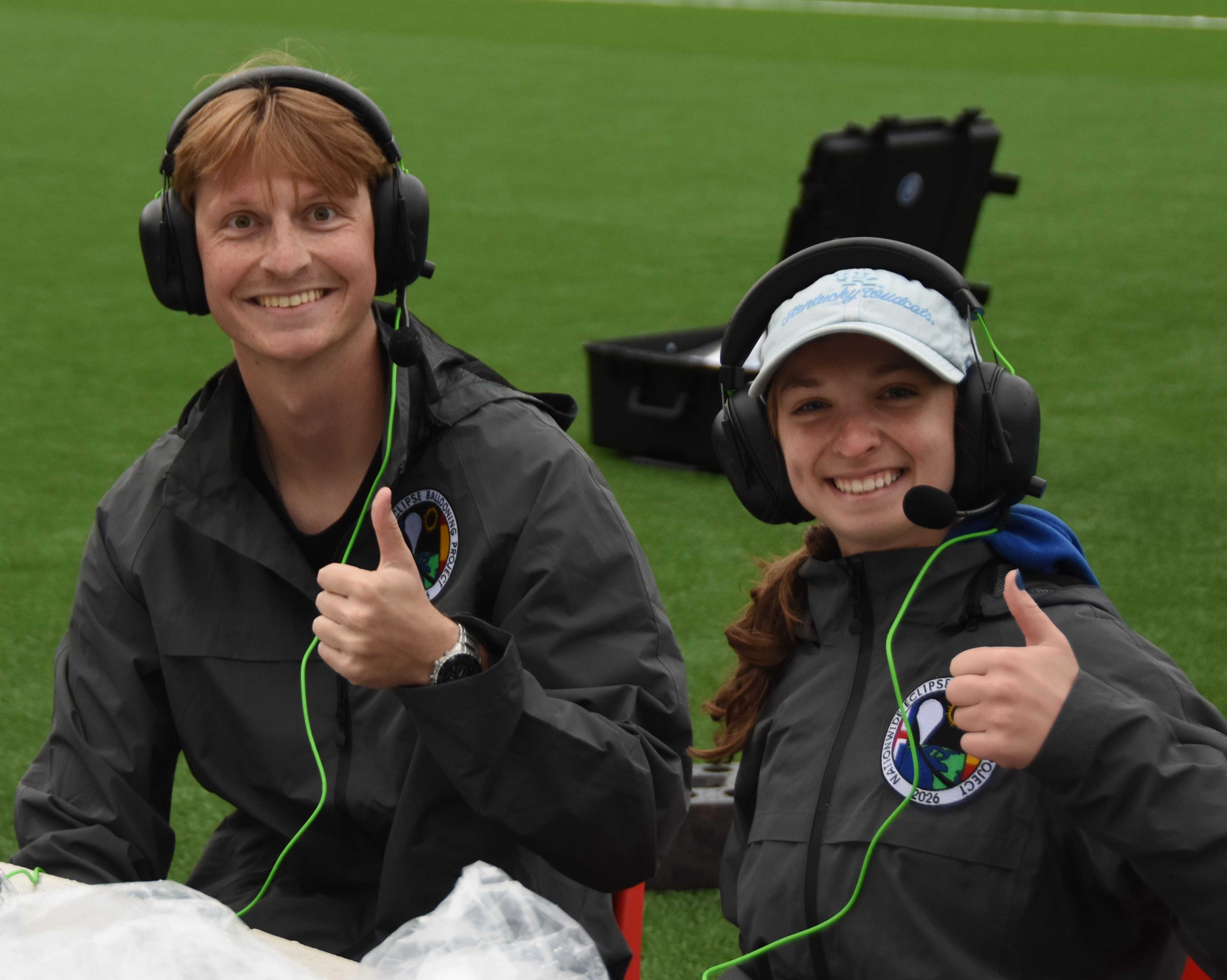 Two students sit at the side of a sports field wearing TV headsets.