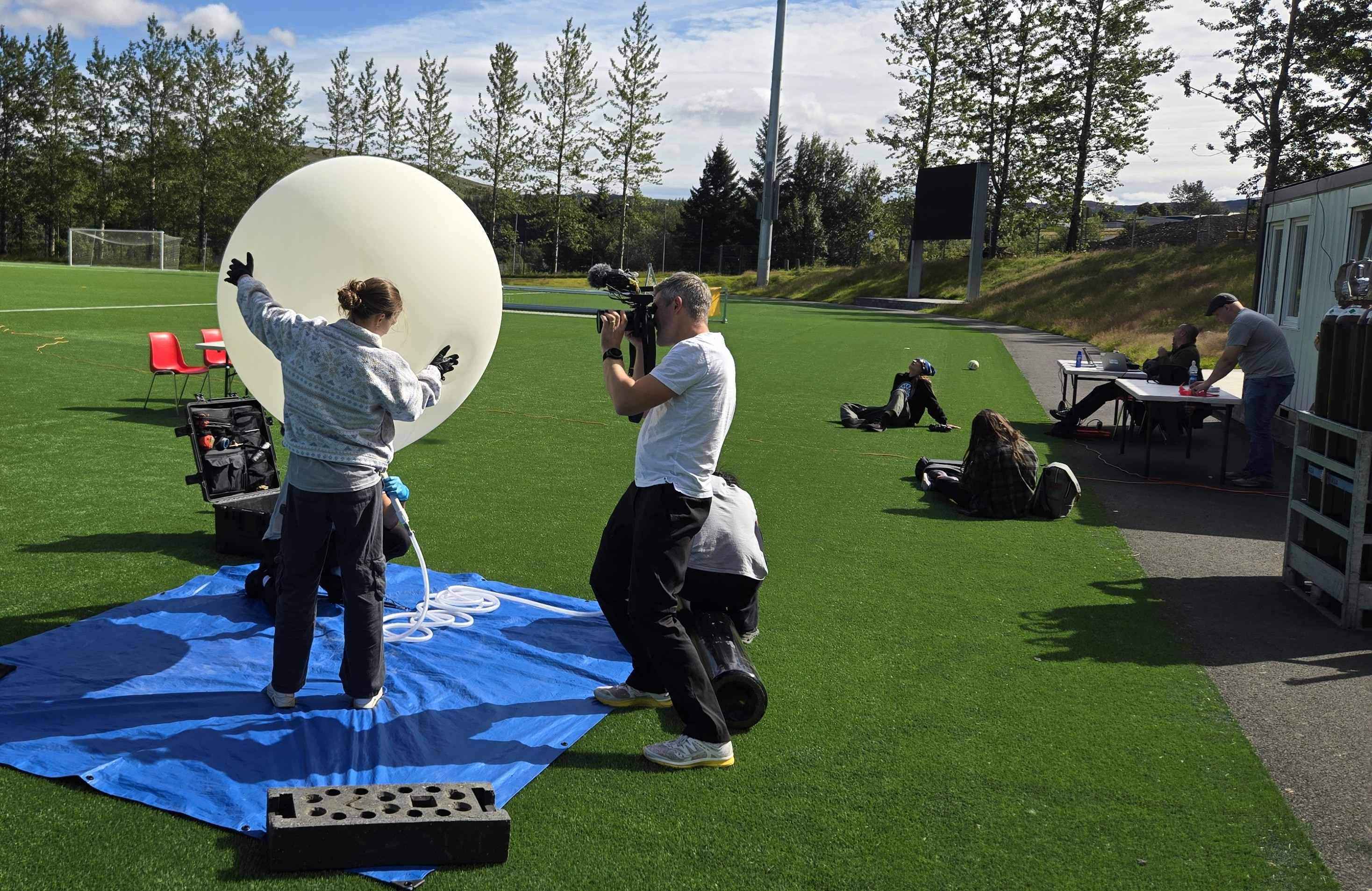 Three students prepare a weather balloon for flight with a media person filming. On the right, more team members prepare equipment ourside a command trailer.