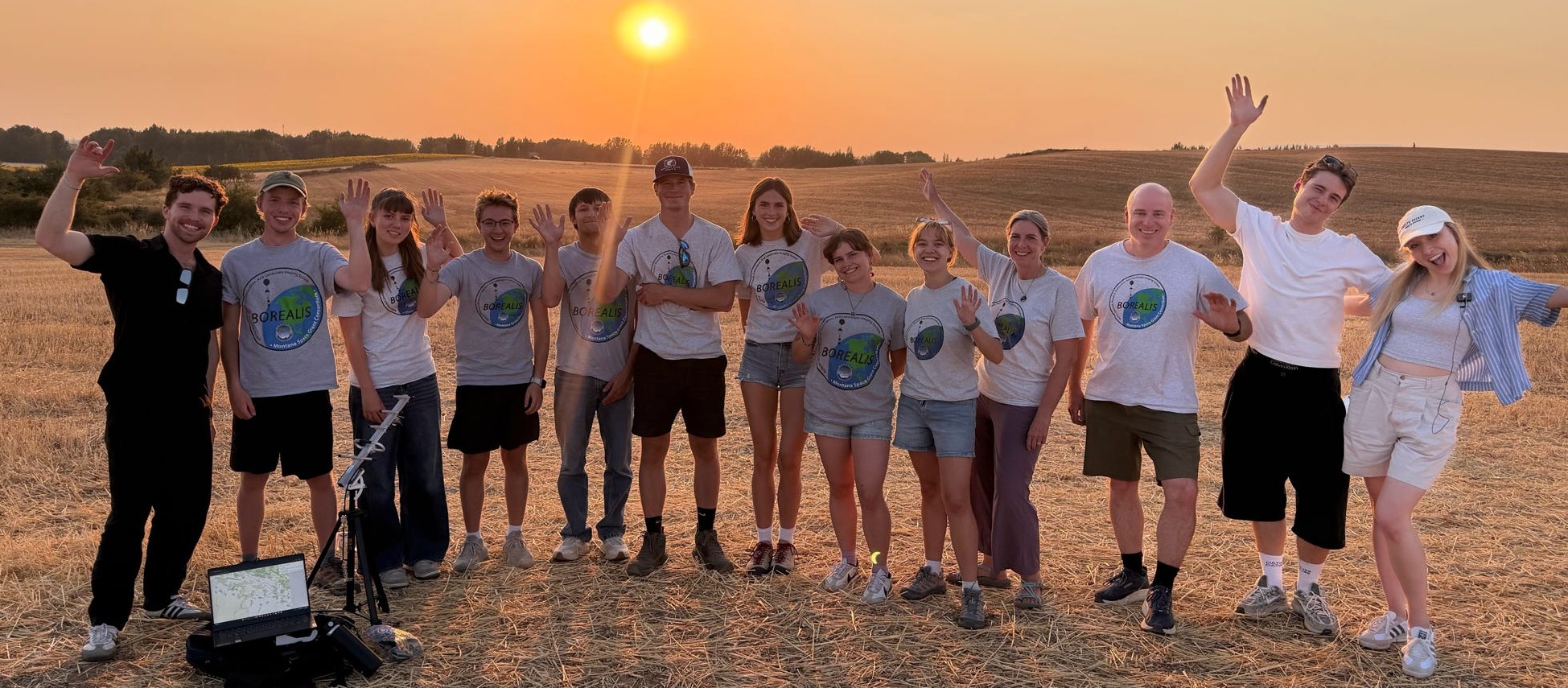 A group of 13 people standing in a field at sunset with happy expressions.