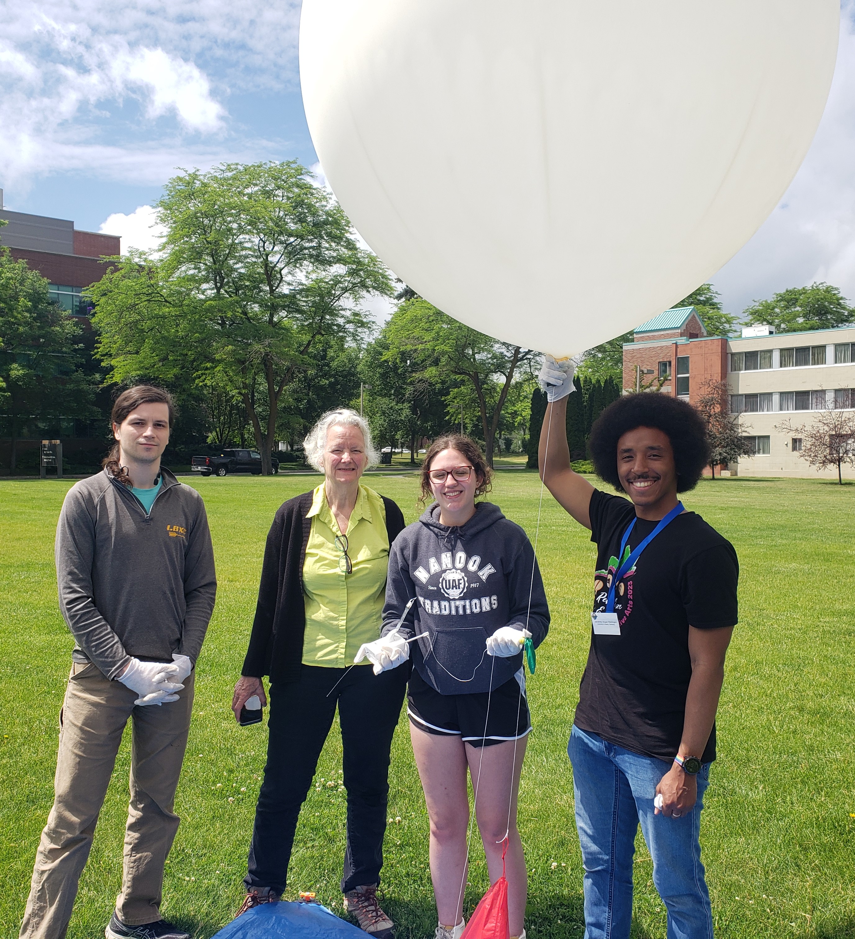 4 people stand preparring to launch a weather balloon that is about 4 feet in diameter with grass and buildings in the background