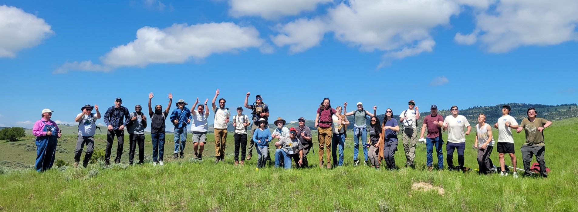 25 people are jumping on a green, grassy hill with mountains and blue sky in the background