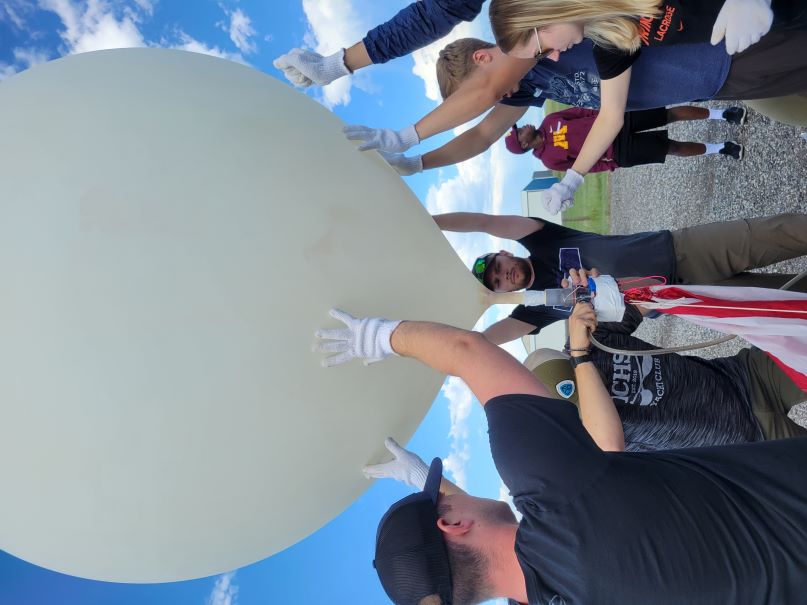 6 people prepare to launch a weather balloon that is about 5 feet in diameter; one student is working on a device just below the balloon; others are wearing gloves so they don't damage the balloon; gravel and blue sky in the background