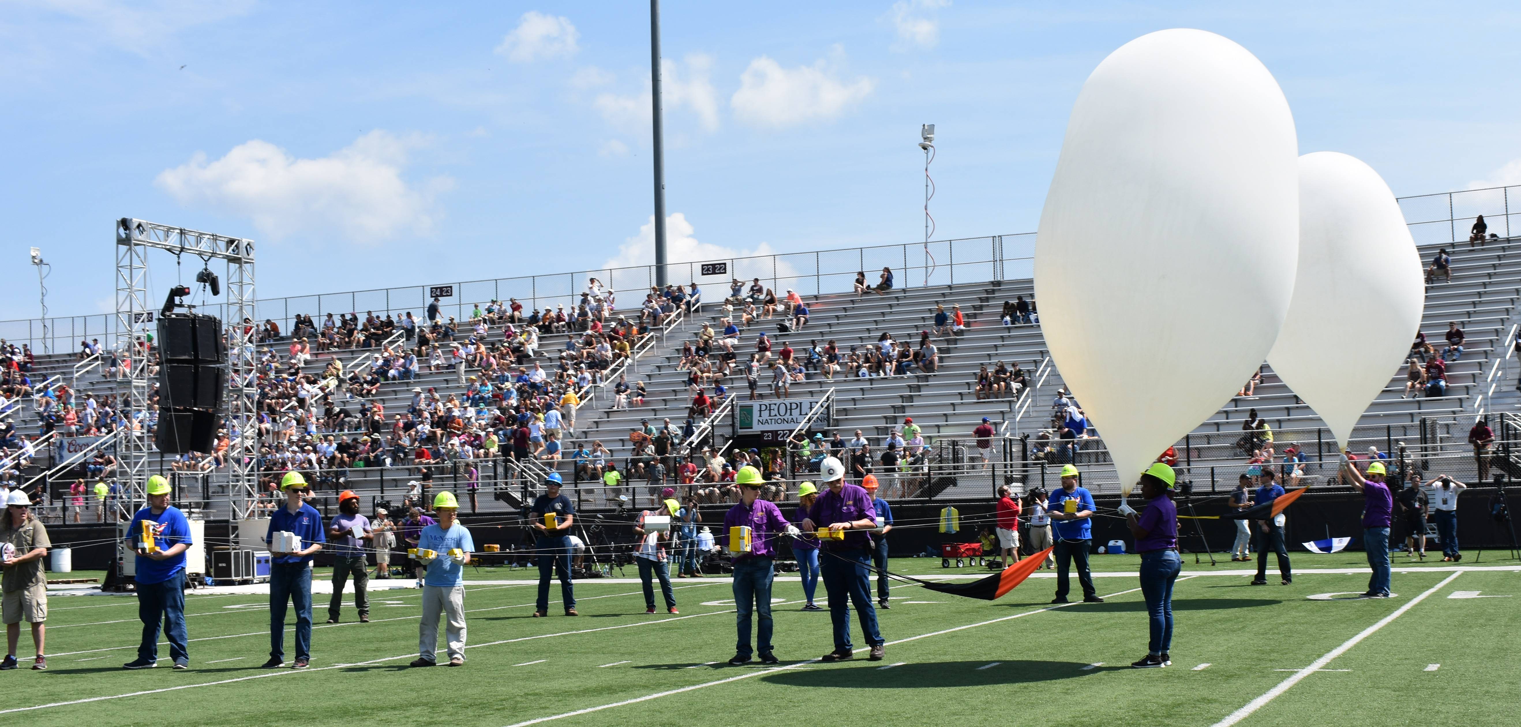 Two balloons about to be released in a football feild with many onlookers in the bleachers