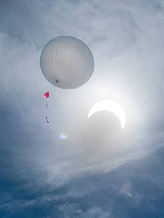 The sky with the sun partially covered by the moon as a balloon is floating up with a string carying payloads