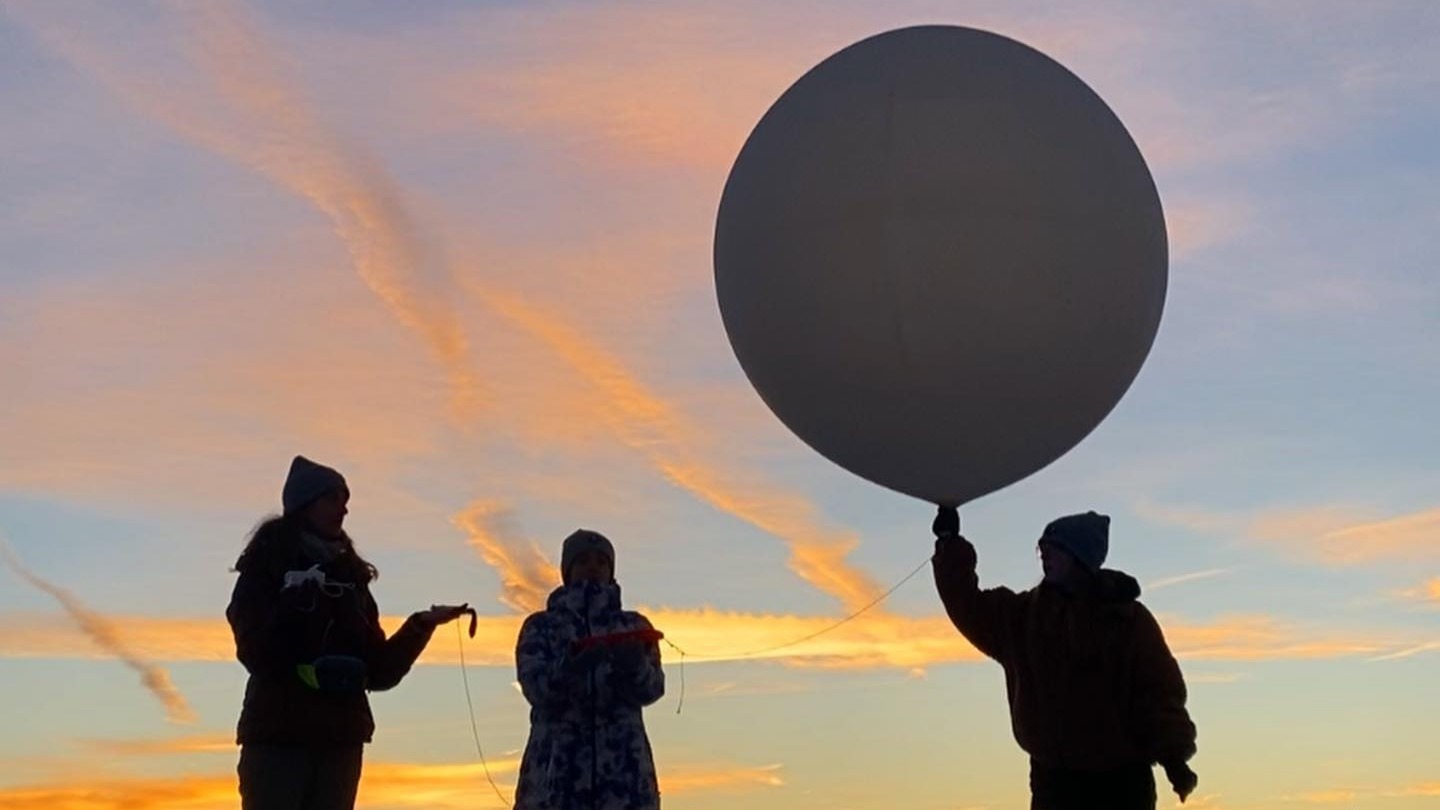 The silhoette of three people holding a balloon against a sunrise.