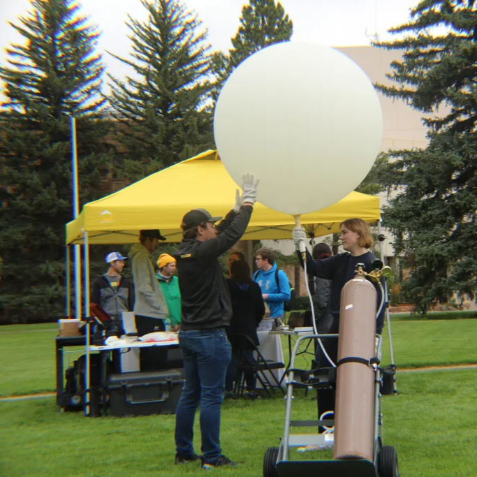 Someone filling a balloon while a team watches under a tent